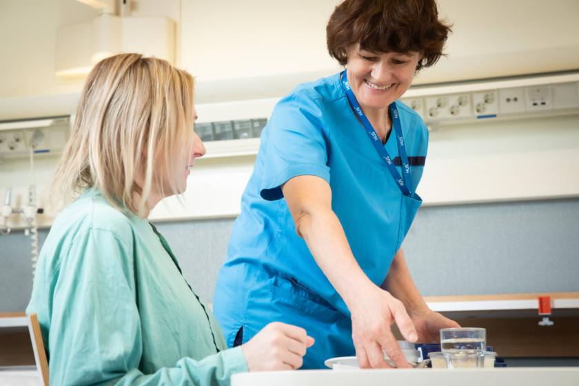 Hymyilevä palvelutyöntekijä tuo istuvalle potilaalle ruoan.