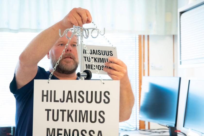 Terveydenhuollon ammattilainen ripustaa lasioveen kylttiä, jossa lukee "Hiljaisuus tutkimus käynnissä".